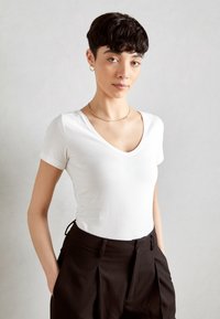 T-shirt blanc ajusté à col en V associé à un pantalon noir taille haute, avec un tissu texturé et des poches latérales. Simple collier en or en accent.