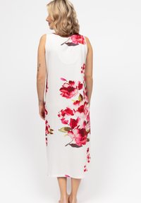 Donna con capelli biondi ondulati che indossa un abito midi bianco senza maniche con grandi stampe floreali rosse e rosa, in piedi a piedi nudi con la schiena rivolta.