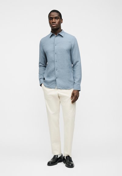 Homme portant une chemise à manches longues bleu clair boutonnée, un pantalon de couleur crème et des chaussures de ville noires, debout devant un fond blanc uni.