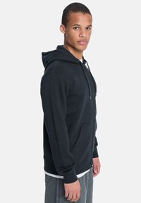 Schwarzer Kapuzenpulli mit Kängurutasche, Kordelzug-Kapuze und gerippten Bündchen. Angenehme Textur und lässige Passform, präsentiert an einem Modell, das seitlich steht.