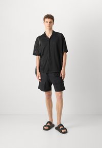 Chemise noire en maille à manches courtes avec boutons, associée à un short cargo noir. La tenue est complétée par des sandales à glissière noires.