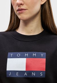Musta huppari, jossa on suorakulmainen graafinen kuva, jossa lukee "TOMMY" ylhäällä ja "JEANS" alla, ja se sisältää punaisia, valkoisia ja sinisiä värejä.