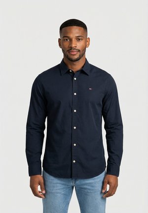 Homme portant une chemise bleu marine ajustée avec des boutons blancs et un jean bleu clair, debout devant un fond blanc uni.