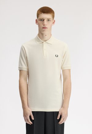 Ung mand med kort rødt hår står vendt mod fronten iført en cremefarvet kortærmet polotrøje med sort logo og sorte bukser.