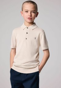 Beige polo tričko s krátkým rukávem a jemnou texturou, s třemi černými knoflíky a malým vyšívaným logem na hrudi, v kombinaci s námořnickými kalhotami.