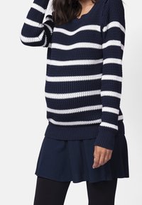 Žena v navy modrém a bílém pruhovaném pleteném svetru přes navy modrou sukni, stojící proti jednoduchému bílému pozadí.