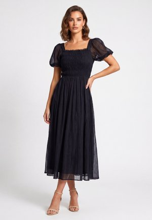 Donna che indossa un vestito midi nero con maniche a sbuffo e scollo quadrato abbinato a tacchi beige con cinturino alla caviglia, in piedi contro uno sfondo semplice.