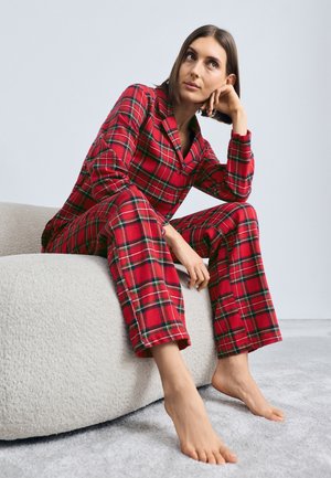 Rødternet pyjamas sæt med en knap-op top og bukser. Blødt stof og afslappet pasform. Model sidder på en tekstureret, lysfarvet sofa.