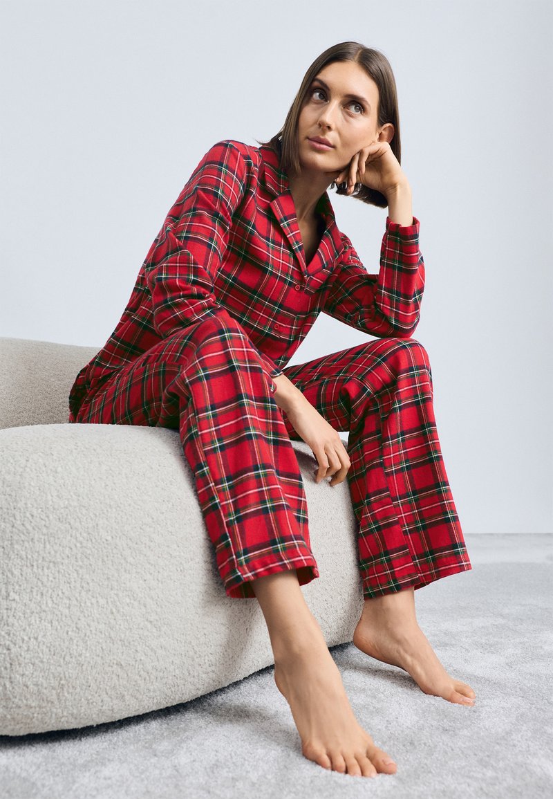 Röd rutigt pyjamasset med en knäppning uppe och byxor. Mjuk tyg och avslappnad passform. Modell sitter på en texturerad, ljus soffa.