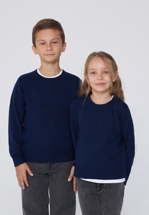 Deux enfants, un garçon et une fille, debout côte à côte portant des pulls bleu marine et des pantalons gris sur un fond clair uni.