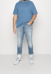 Camiseta de algodón azul con mangas cortas y logo, combinada con jeans ajustados de mezclilla claros y zapatillas blancas con detalles grises.