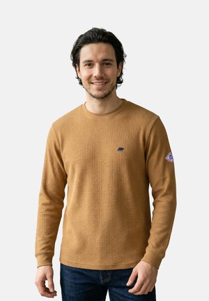 Mann mit dunklen Haaren, der ein beigefarbenes, strukturiertes Langarmshirt mit kleinem Bärenlogo und einem Aufnäher am linken Ärmel trägt, steht und lächelt.