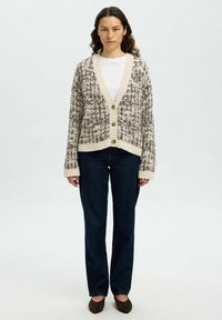 Strickjacke in Creme und Braun mit V-Ausschnitt, Knopfverschluss, strukturiertem Muster und gerippten Bündchen, kombiniert mit dunklen Jeans und braunen Schuhen.