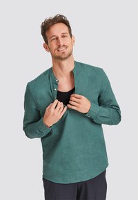 Green, long-sleeve shirt with a mandarin collar, featuring a button placket and a soft texture. Worn open over a black undershirt.