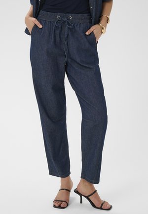 Kvinde iført mørkeblå denim joggerbukser med elastisk talje og snøre, kombineret med sorte hælede sandaler og gyldne armbånd.