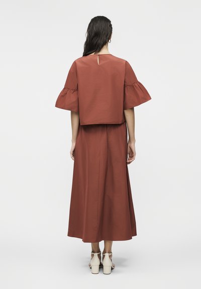 Γυναίκα με μακριά σκούρα μαλλιά, φοράει φόρεμα σε χρώμα σκουριάς με φαρδιές ριγωτές μανσέτες και κρεμ σανδάλια με χοντρό τακούνι, στέκεται γυρισμένη προς τα πίσω.