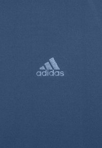 Blå atletisk t-shirt med en texturerad, reflekterande vit Adidas-logga på vänster sida. Materialet verkar slätt och stretchy.