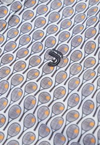 Witte stof met een herhaald patroon van blauwe tennisrackets en oranje tennisballen, met een zwart geborduurd logo erbovenop.