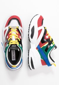 Zapatillas coloridas que presentan una mezcla de materiales rojos, negros, blancos y verdes. Incluyen cordones amarillos y una marca prominente en la lengüeta.