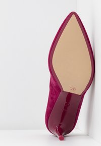 Chaussure à talon haut en daim fuchsia à bout pointu avec talon stilettos brillant, taille 37 indiquée sous la semelle, placée debout contre un mur d'angle blanc.