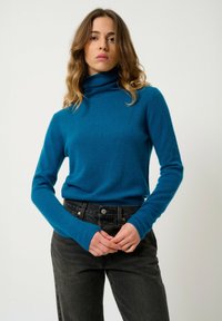 Türkiser Rollkragenpullover aus weichem, strukturiertem Stoff; enganliegende lange Ärmel und eine lässige Silhouette, kombiniert mit dunklen, hochgeschnittenen Jeans.