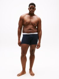 Uomo adulto in piedi scalzo su uno sfondo bianco, indossa boxer blu navy con vita bianca.