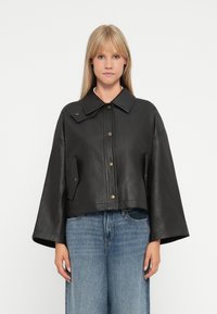 Veste en cuir noir courte avec un large col, fermeture avant par boutons et poches sur les côtés. Texture lisse et manches longues ample.
