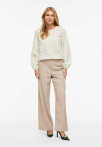 Femme aux longs cheveux blonds portant un chemisier blanc à manches longues brodé, un pantalon beige à jambes larges et des talons noirs avec des détails en chaîne dorée.