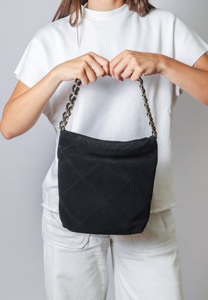 Femme en blanc tenant un sac à main matelassé noir avec une bandoulière chaîne dorée par ses anses sur un fond neutre.