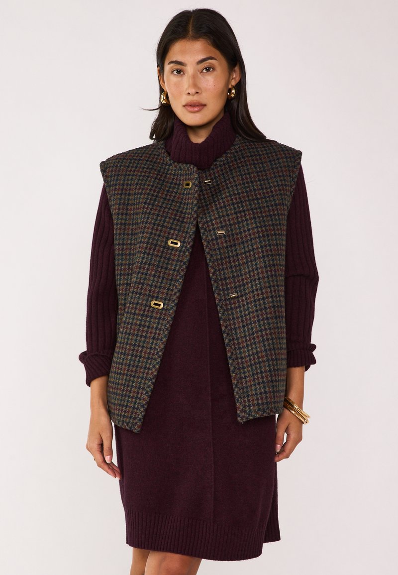Gilet in tweed di lana nei toni del verde scuro, marrone e blu, con collo alto e dettagli in oro; indossato sopra un vestito bordeaux.