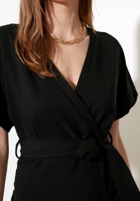 Robe noire en tissu fluide avec des manches courtes, mettant en avant un détail de taille nouée et un décolleté en V profond. Collier en chaîne dorée en accent.