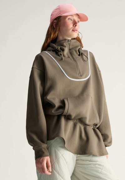 Žena v oversized olivovém svetru s přední kapsou a zipem, růžové kšiltovce a bílých prošívaných kalhotách, dívající se do strany.