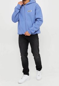 Jasnoniebieska bluza z zamkiem, wykonana z miękkiego materiału, z kieszenią z przodu i naszywką z logo. Połączona z czarnymi dżinsami i białymi trampkami.