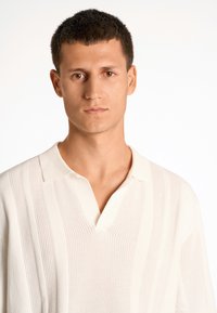 Pletené polo tričko v off-white farbe s vertikálnymi pruhmi, krátkymi rukávmi a rozdeleným golierom. Hladká textúra a voľný strih.