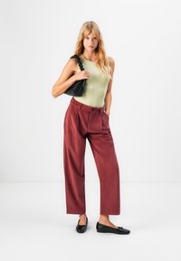 Donna con un top senza maniche color oliva, pantaloni a vita alta in velluto a coste ruggine a gamba larga, ballerine nere e una borsa a spalla arricciata nera.