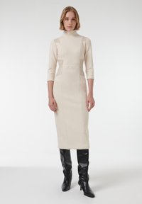 Beige gebreide jurk met een hoge kraag, 3/4 mouwen en details aan de zijkanten, gecombineerd met zwarte kniehoogte laarzen met hak. Eenvoudige, aangesloten silhouet.