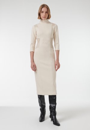 Vestido de punto beige con cuello alto, mangas 3/4 y detalles en los paneles laterales, combinado con botas de tacón negro hasta la rodilla. Silueta simple y ajustada.