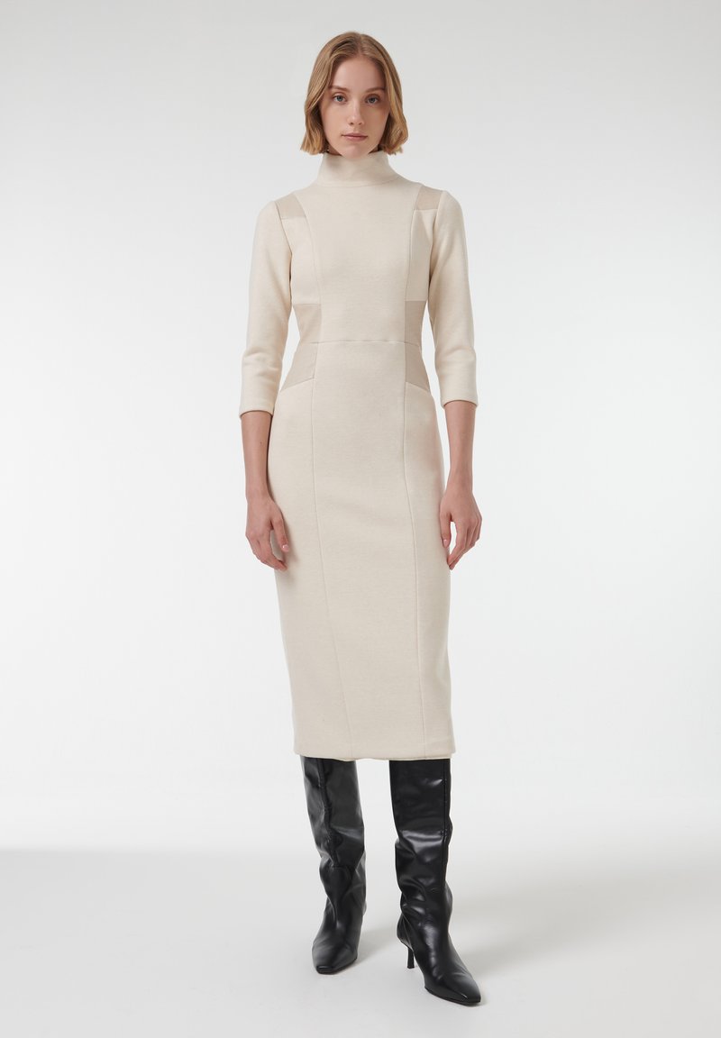 Beige gebreide jurk met een hoge kraag, 3/4 mouwen en details aan de zijkanten, gecombineerd met zwarte kniehoogte laarzen met hak. Eenvoudige, aangesloten silhouet.