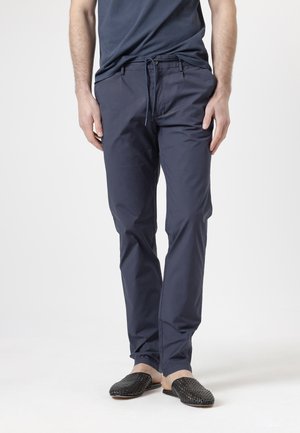 Hombre con pantalones azul marino con cordón, zapatos negros tejidos sin cordones y camisa azul marino, de pie frente a un fondo blanco liso.