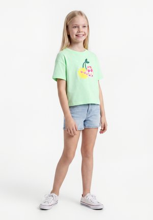 Fille souriante aux longs cheveux blonds porte un T-shirt vert clair avec un motif de fruit, un short en jean et des baskets blanches, debout sur un fond blanc.