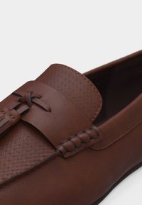 Καφέ δερμάτινο μοκασίνι με υφή στην επάνω επιφάνεια, διακοσμητικό δερμάτινο κόμπο και πλεκτή λεπτομέρεια περιμετρικά, σε ανοιχτό φόντο.