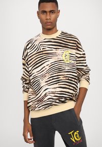 Beige och svart randig sweatshirt med en ribbad krämfärgad krage, prydd med en gul logotypdetalj. Buren med mörka byxor, vilket visar en avslappnad passform.