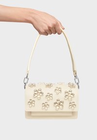 Sac à main couleur crème avec des ornements floraux et une texture lisse. Comprend une poignée supérieure et des accents de ferrures métalliques.