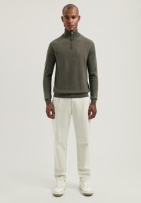 Donkergroene zip-neksweater, geribbelde mouwen, gecombineerd met witte broek en witte sneakers. Eenvoudig, casual ontwerp met een gestructureerde afwerking.