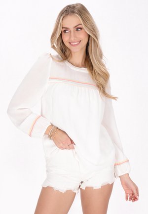 Witte langslief top met textuur en geborduurde accenten in het roze, gecombineerd met versleten witte shorts. Casual outfit.