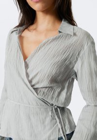 Haut en wrap gris clair en tissu texturé, avec un col en V, des manches longues et une taille ceinturée avec un détail de lien.