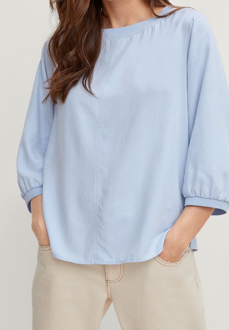 Femme portant un chemisier bleu clair à manches longues avec poignets froncés et un pantalon beige, les mains posées négligemment dans les poches, sur un fond uni.