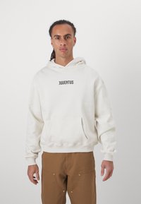 Fehér kapucnis sweatshirt, amelyen fekete színnel van nyomva a "JUVENTUS" felirat, kanguru zsebbe és bordázott alsó szegélye van. Barna nadrággal párosítva, egyszínű és texturált anyagból.