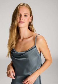 Satin-graues Kleid mit drapiertem Ausschnitt und schmalen Trägern, das eine glatte Textur und eine lockere Passform aufweist. Das Model posiert mit den Händen an der Taille.