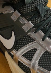 Sportskor med ovandel i grått nät och gröna accenter, med en vit logotyp. Har strukturerade overlay och snörningsdesign för stöd.